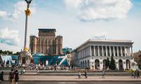Photograph of Maidan square in Kiyv, Ukraine. The image shows buildings, a monument column and people walking the streets 